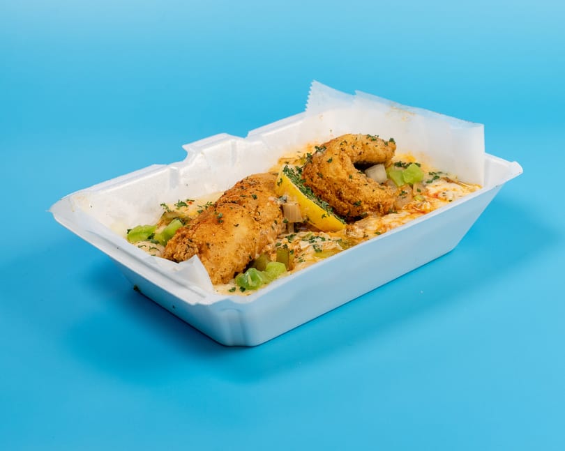 Fried fish fillets with sauce, green peppers, and a lemon wedge in a white takeout container.