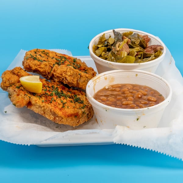 Fried fish with a lemon wedge, collard greens, and baked beans in white containers on a blue background.
