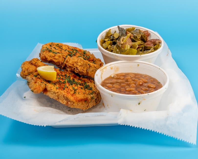 Fried fish with a lemon wedge, collard greens, and baked beans in white containers on a blue background.