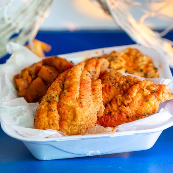 Fried fish fillets and sweet potato chunks in a white takeout container on a blue surface.