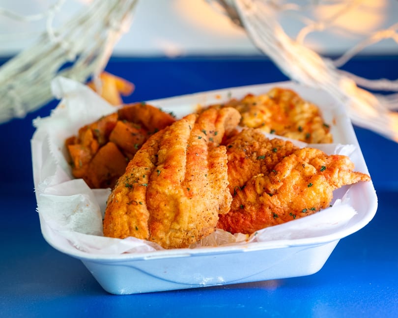 Fried fish fillets and sweet potato chunks in a white takeout container on a blue surface.