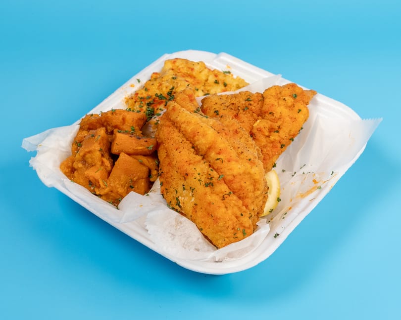 Fried fish fillets, sweet potatoes, and rice in a white takeout container on a blue background.