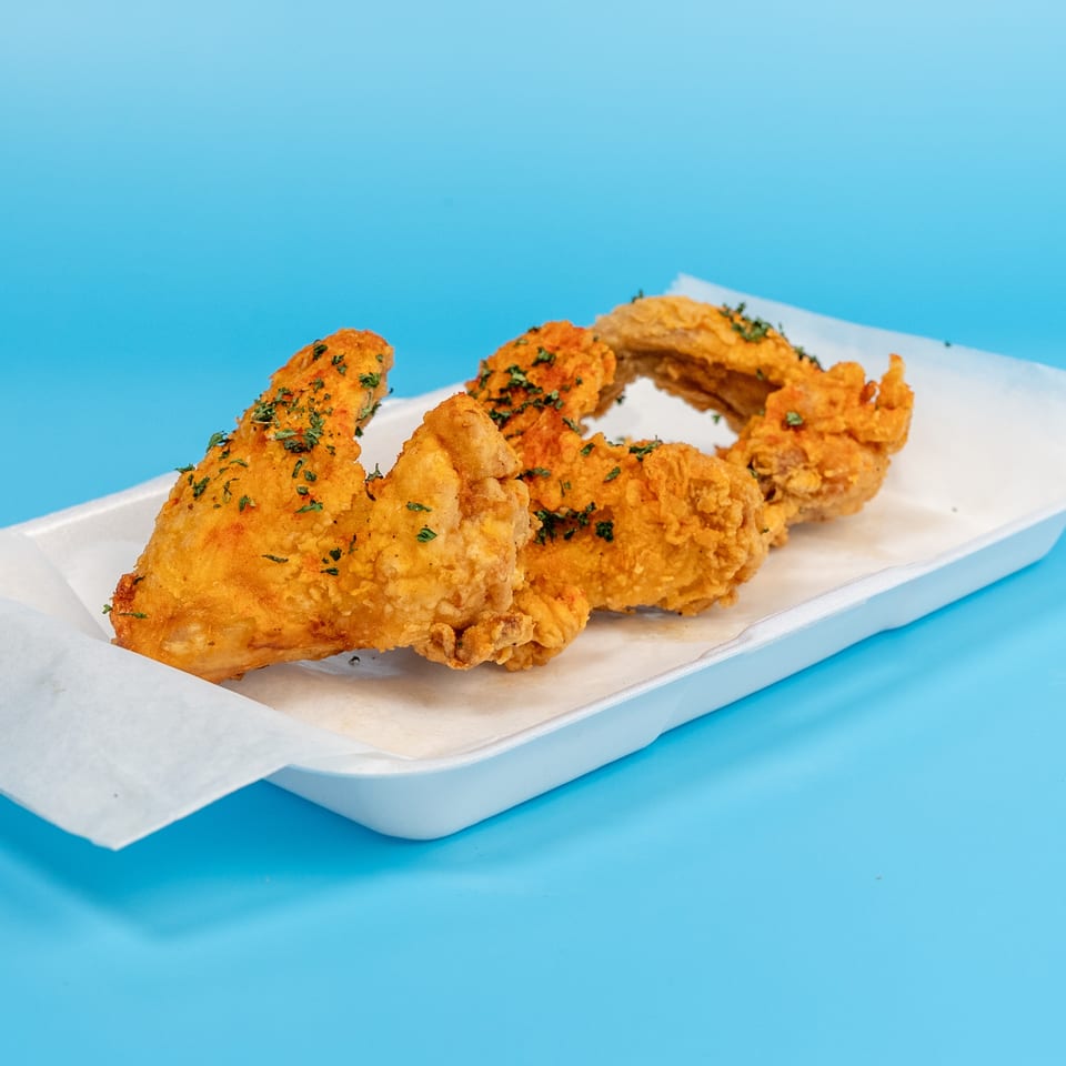 Four golden-brown fried chicken wings with herbs in a white tray on a blue background.
