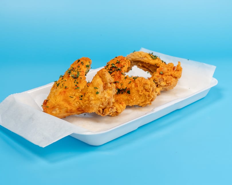 Four golden-brown fried chicken wings with herbs in a white tray on a blue background.