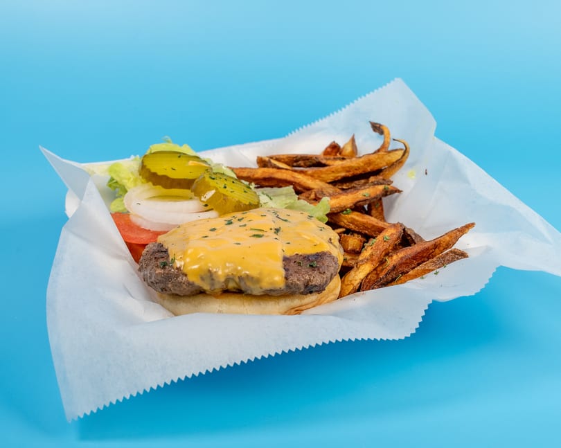 Cheeseburger with lettuce, tomato, onion, pickles, and a side of fries in a paper-lined basket.