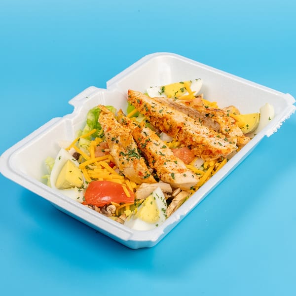 Chicken salad with lettuce, cheese, tomatoes, mushrooms, and hard-boiled eggs in a takeout container.