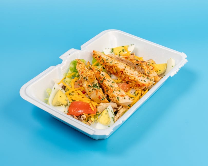 Chicken salad with lettuce, cheese, tomatoes, mushrooms, and hard-boiled eggs in a takeout container.